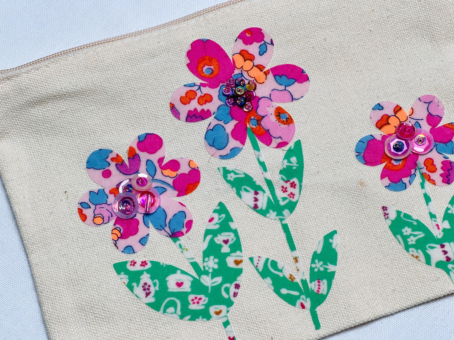 May include: A beige canvas zippered pouch featuring three floral appliques. Each flower has pink, blue, and orange patterned petals with a cluster of shiny pink and purple sequins at the center. The stems and leaves are green with a white teapot pattern.