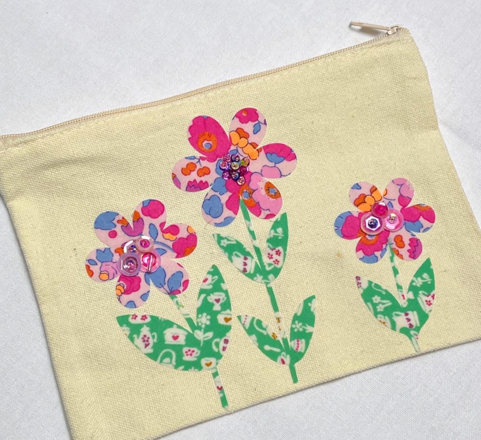May include: A rectangular cream-colored canvas zipper pouch featuring three floral appliqués. Each flower has patterned pink, blue, and orange petals with green stems and leaves. The centers of the flowers are embellished with small, shiny pink sequins.