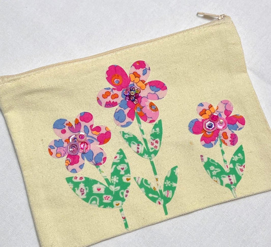May include: A rectangular cream-colored canvas zipper pouch featuring three floral appliqués. Each flower has patterned pink, blue, and orange petals with green stems and leaves. The centers of the flowers are embellished with small, shiny pink sequins.